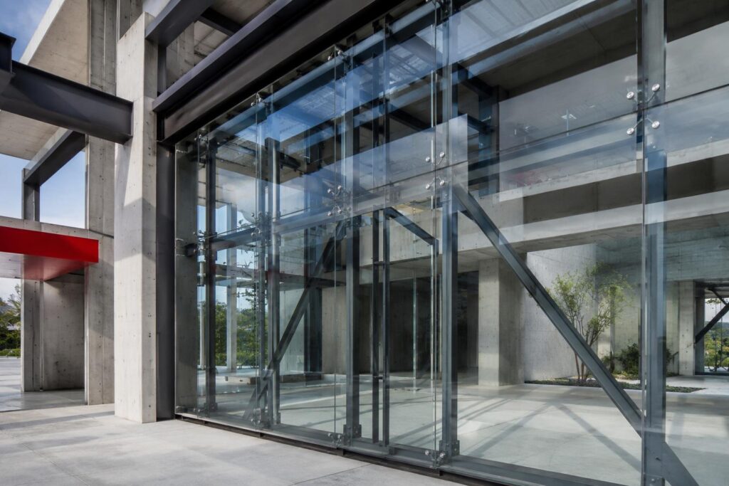 Modern structural glass facade with transparent panels, steel framing, concrete supports, and visible glass hardware
